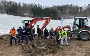 Symbolischer Spatenstich für Glasfaserverlegung mit Vertretern und Bagger im Hintergrund
