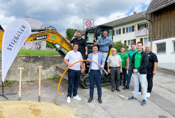 Das Stiegeler Netzbauteam und Vertreter der Tiefbaufirma zusammen mit dem Hilzinger Bürgermeister Holger Mayer und Mitgliedern des Ortschaftsrats vor einem Bagger und Erdhaufen beim Spatenstich für den Glasfaserausbau in Binningen.