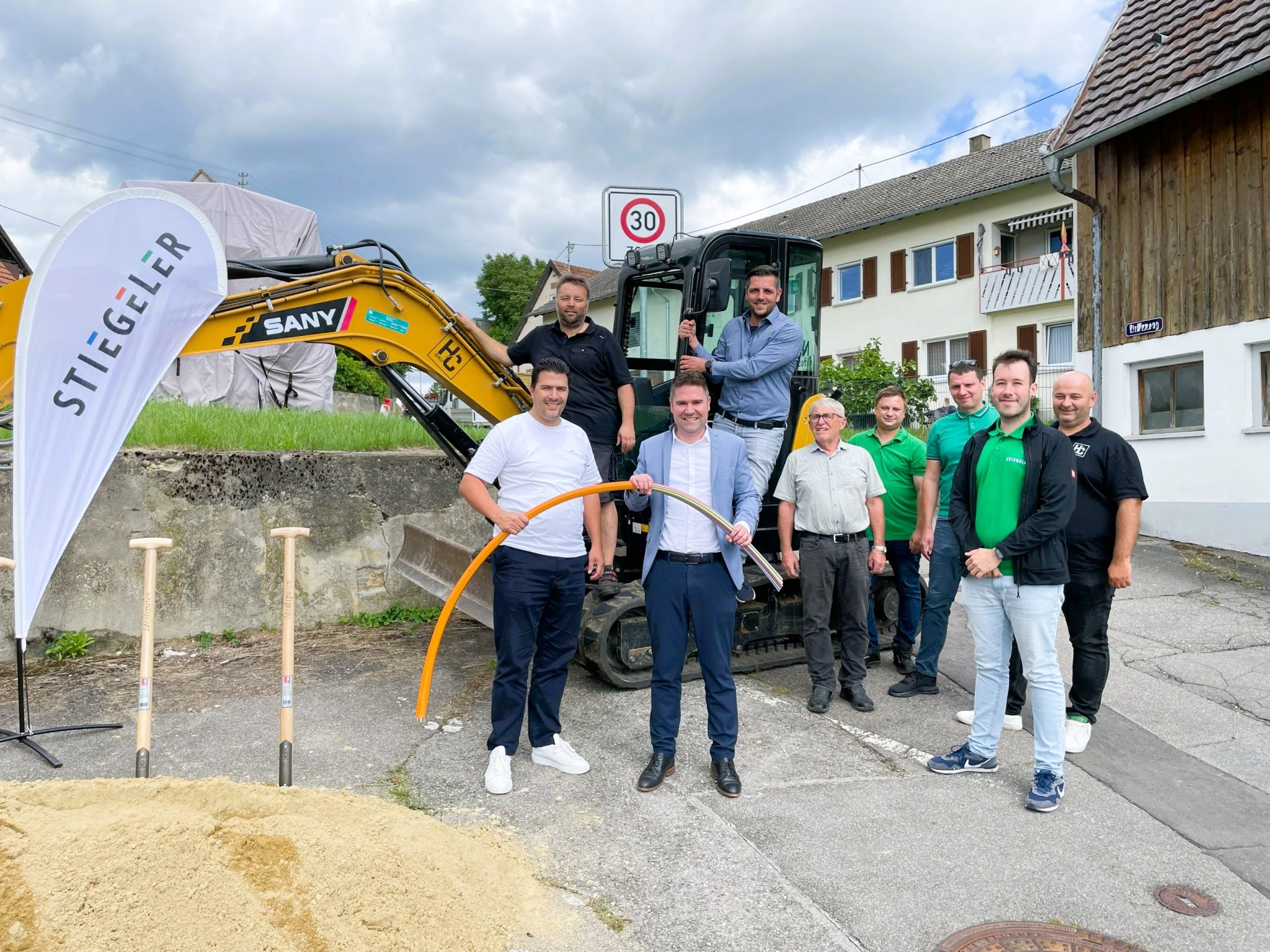 Das Stiegeler Netzbauteam und Vertreter der Tiefbaufirma zusammen mit dem Hilzinger Bürgermeister Holger Mayer und Mitgliedern des Ortschaftsrats vor einem Bagger und Erdhaufen beim Spatenstich für den Glasfaserausbau in Binningen.