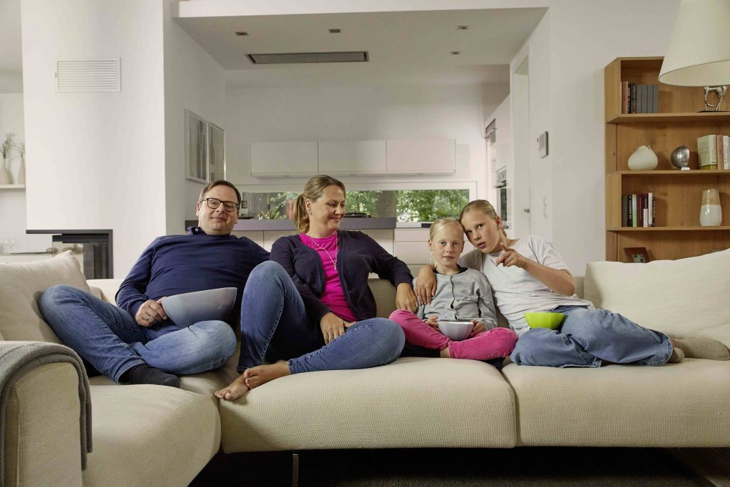 Ein Bild aus der neuen Stiegeler-Herbstkampagne: Eine Familie mit Vater, Mutter und zwei Kindern sitzt entspannt mit Popcorn auf einem Sofa in einer modern eingerichteten Wohnung. In der neuen Stiegeler-Herbstkampagne „Damit Märchen glücklich enden“ verfolgt die Familie die Geschichte von Hänsel und Gretel auf dem heimischen Fernseher.